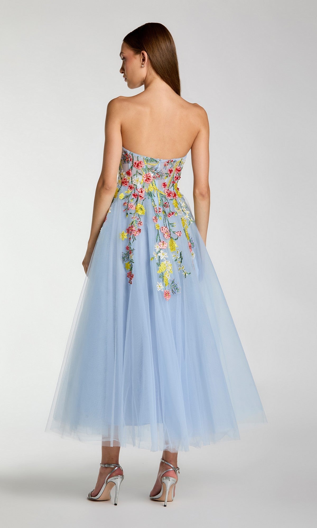 A woman stands with her back to the camera, wearing the Mac Duggal 21304 Tea-Length Party Dress—a strapless, light blue tulle gown adorned with colorful floral embroidery on the back.